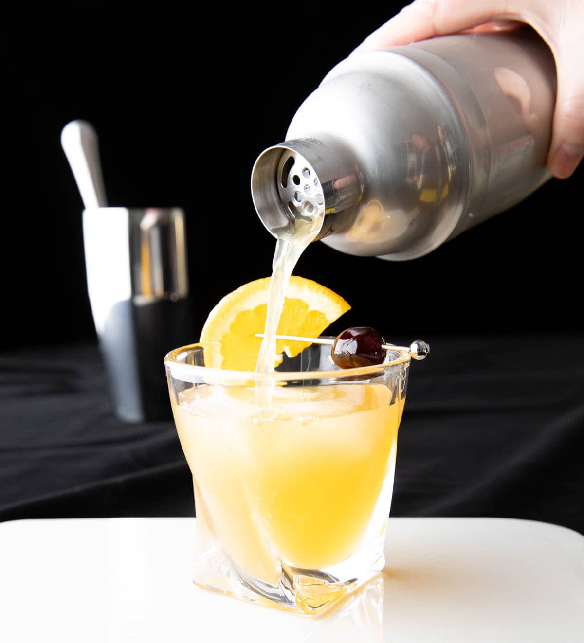 A photo showing How to Make a Whiskey Sour – pouring the last drop over the glass with an orange and cherry garnish