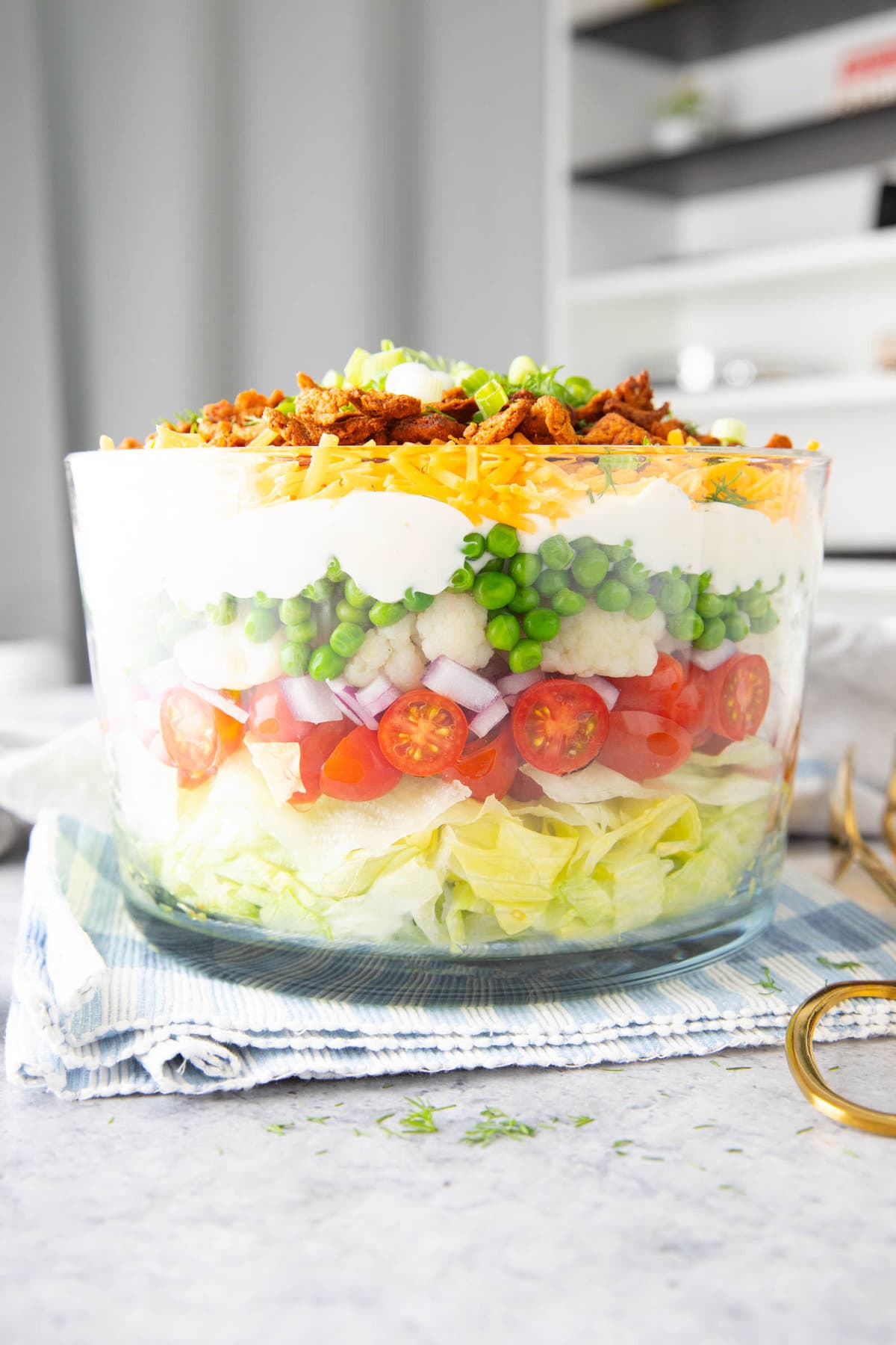 7 Layer Salad prepared in a trifle glass bowl with layers of tomatoes, peas, onions, dressing and more.