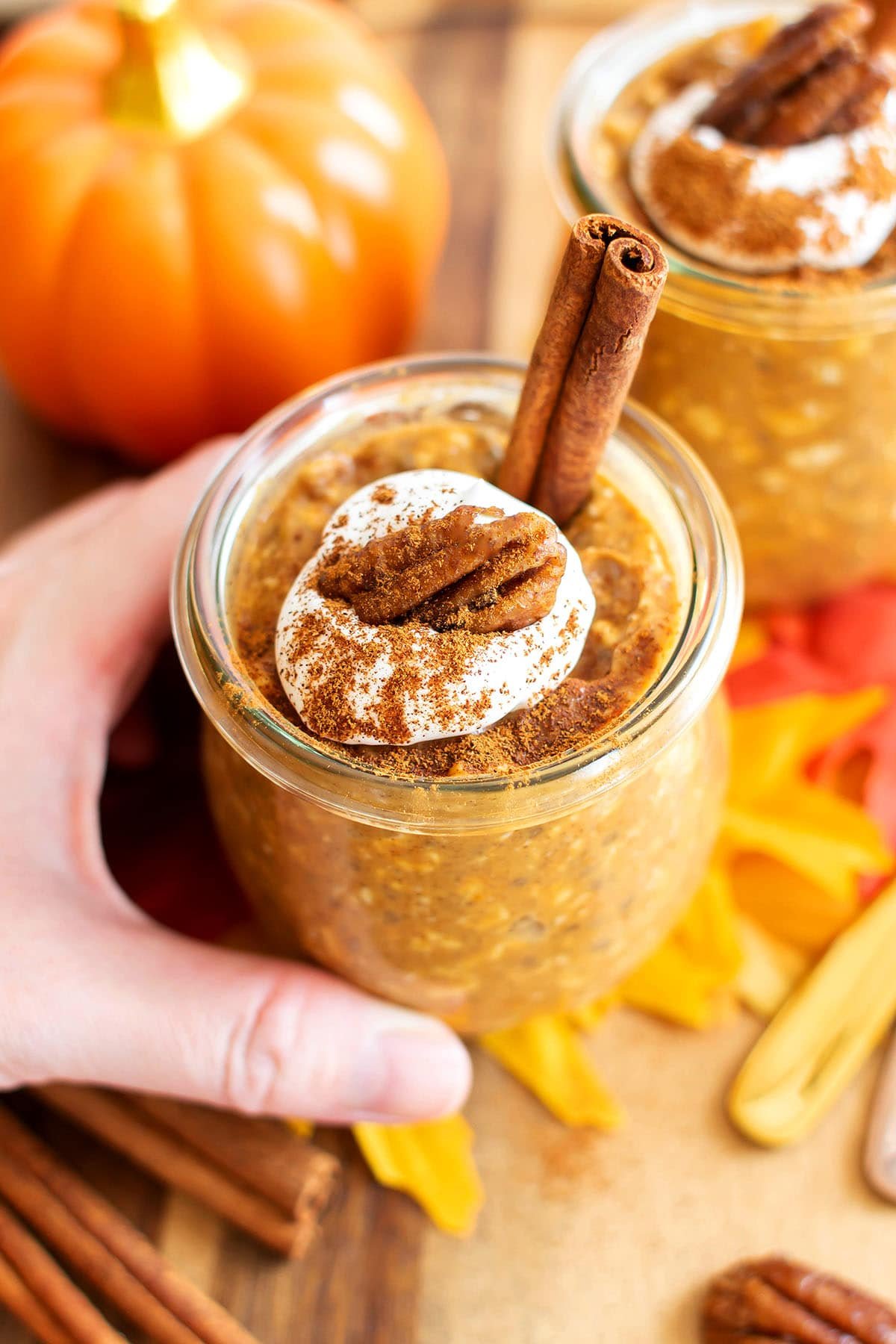 Someone holding a jar of pumpkin chia overnight oats topped with a cinnamon stick and whipped cream with a pecan