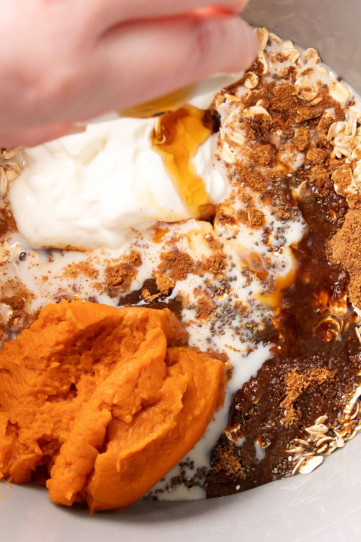 Pumpkin pie overnight oats ingredients in a mixing bowl including chia seeds, rolled oats, cinnamon, pumpkin pie spice, pumpkin puree and someone adding maple syrup