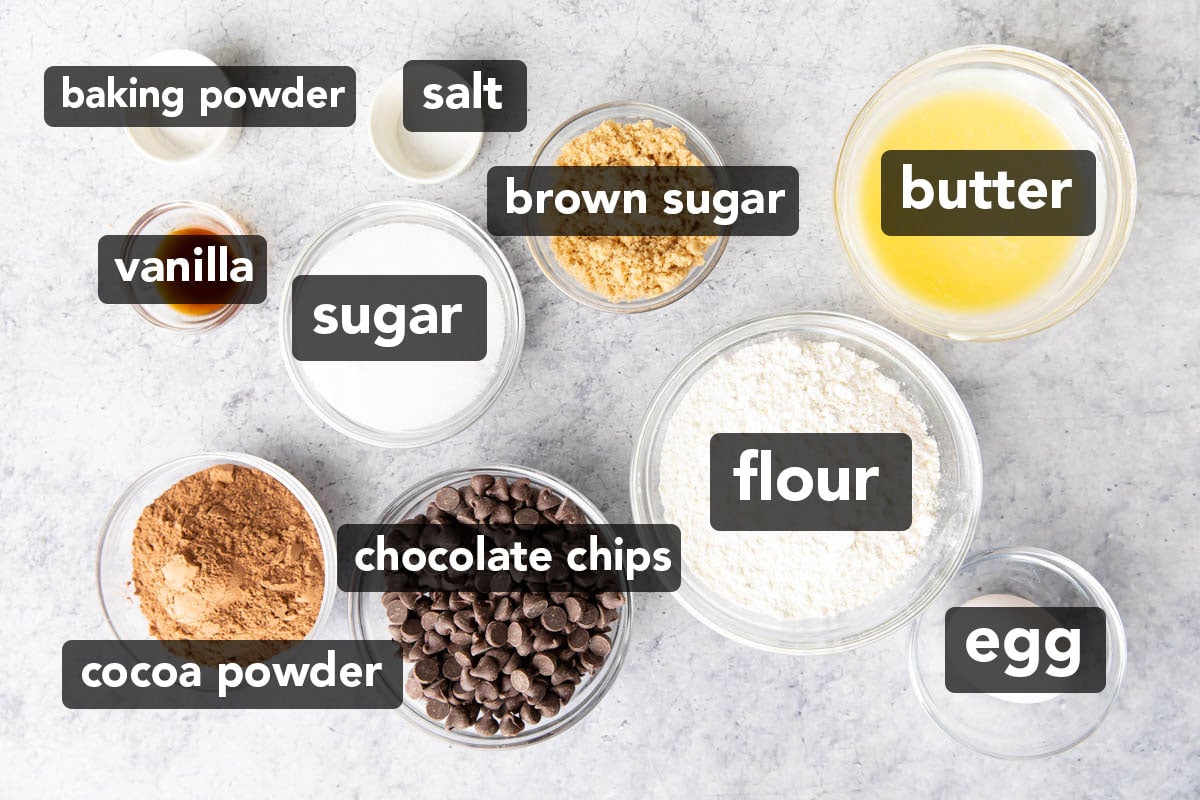 Ingredients to make Brownie Cookies laid out in bowls, including cocoa powder, brown sugar, granulated sugar, flour, egg, butter, baking powder, vanilla, and chocolate chips.