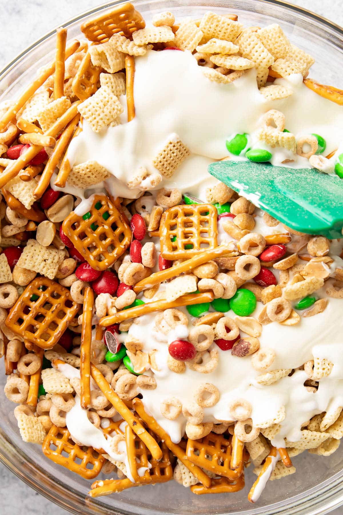 Close up of green spatula stirring white chocolate throughout bowl of chex mix, pretzels, M&M’s and more to coat