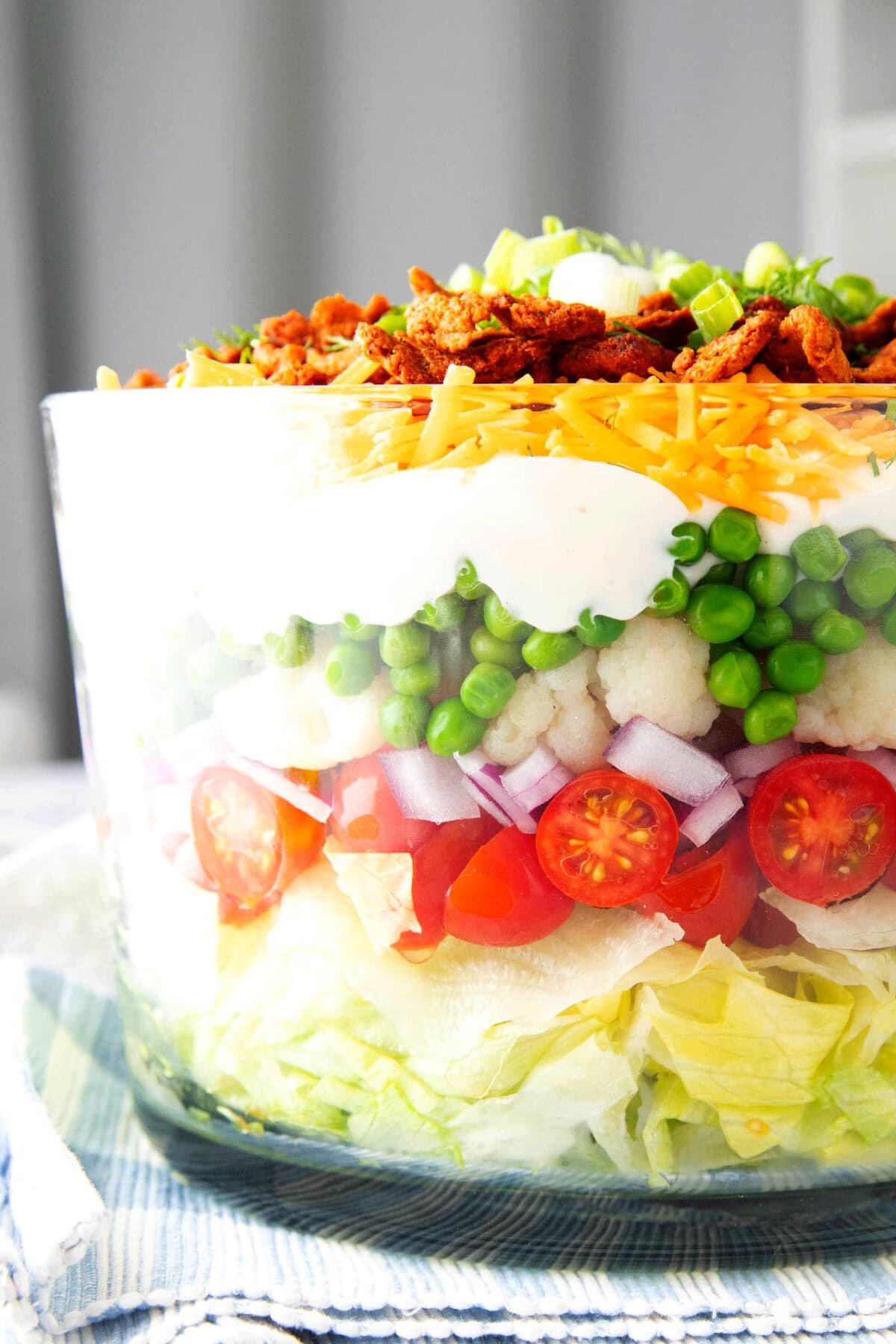 Close up of this traditional 7 layer salad recipe shows cheddar cheese, bacon, and green onions on top of dressing and layers of peas, cauliflower, red onions, grape tomatoes, and lettuce.