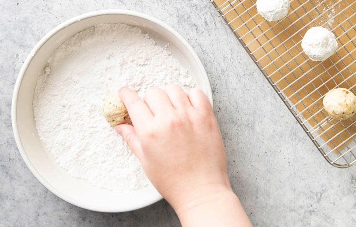 Rolling baked snowball cookies in a rimmed bowl of powdered sugar