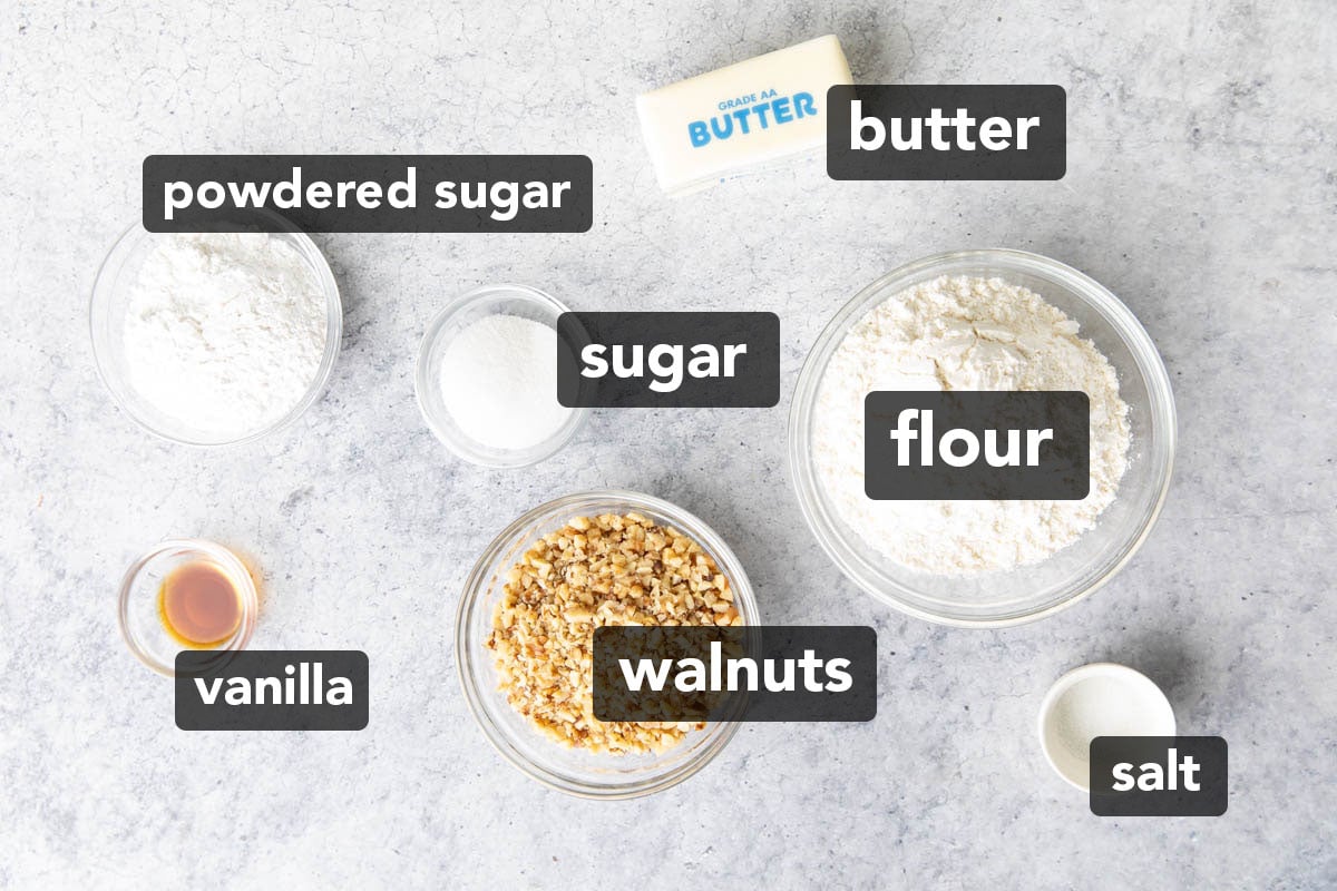 Ingredients in the recipe for snowball cookies measured into glass bowls including chopped walnuts, flour, butter, sugar, and powdered sugar