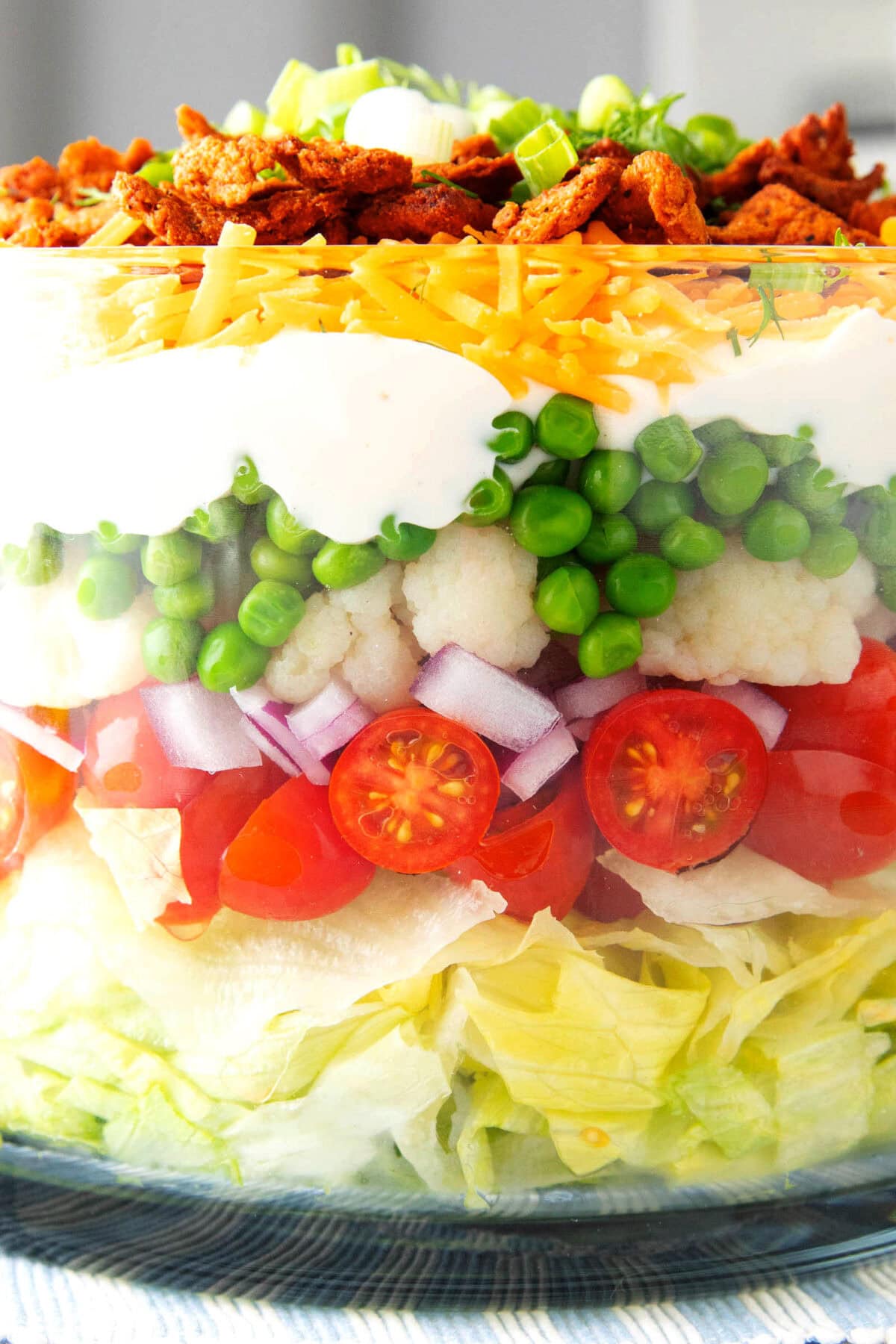 Close up of Seven Layer salad to showcase layers, including peas, cauliflower, red onions, tomatoes, mayonnaise dressing, cheese, and bacon.