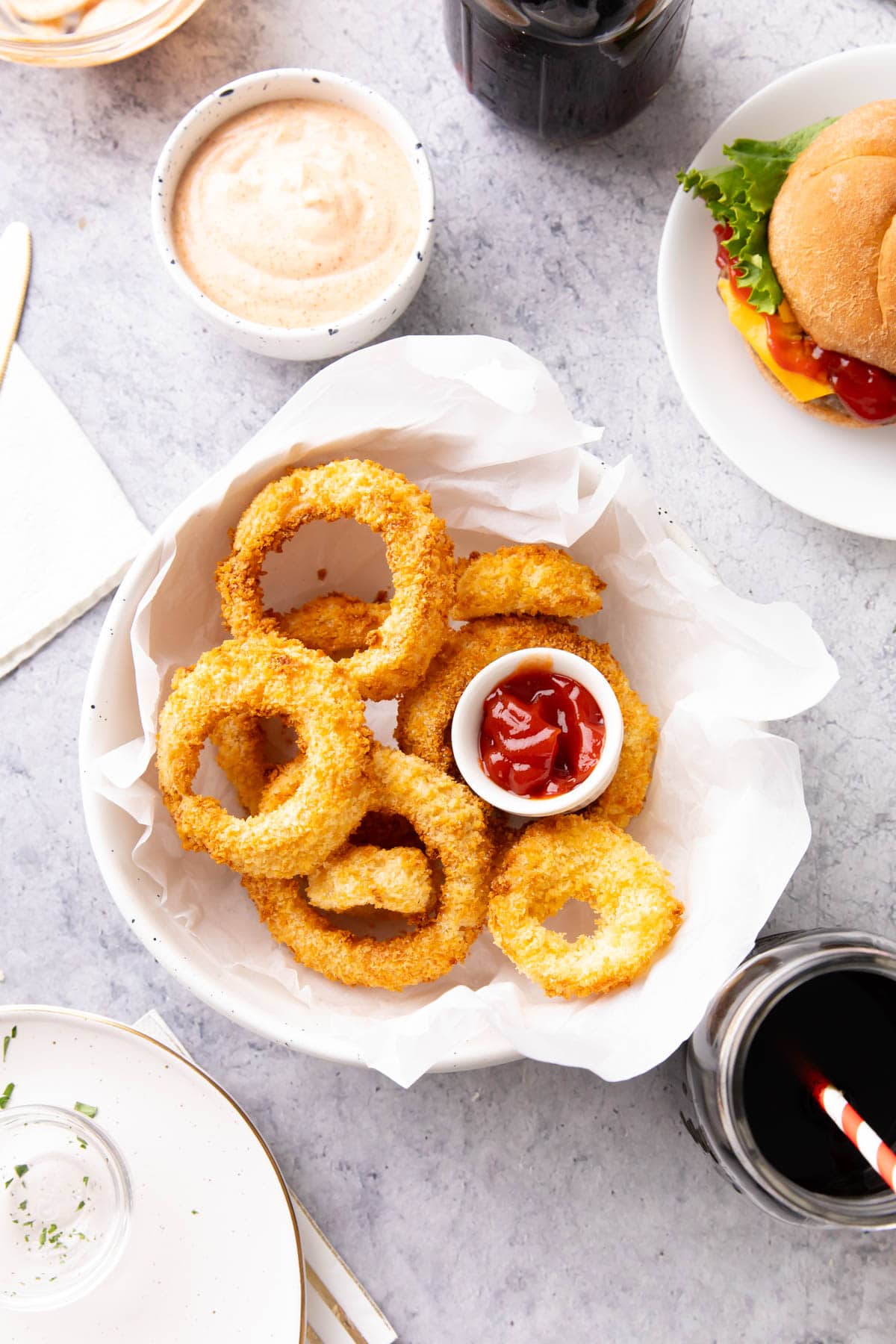 A diner table set up with a bowl of crispy, crunchy air fryer onion rings, onion ring sauce, ketchup, a burger, and glasses of soda.