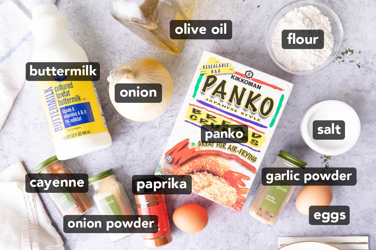 Ingredients for making Onion Rings in an Air Fryer on a table, including Panko bread crumbs, buttermilk, a medium sweet onion, eggs, flour, paprika, onion powder, garlic powder, oil, and salt.