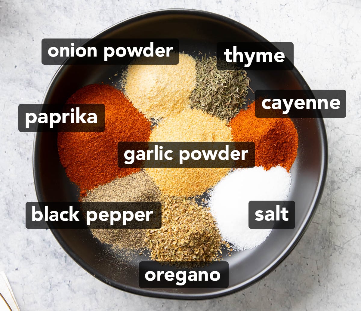 Ingredients for this Cajun Seasoning Recipe sitting in neat piles in a rimmed black dish including smoked paprika, garlic powder, salt, ground black pepper, onion powder, dried oregano, cayenne, and dried thyme.