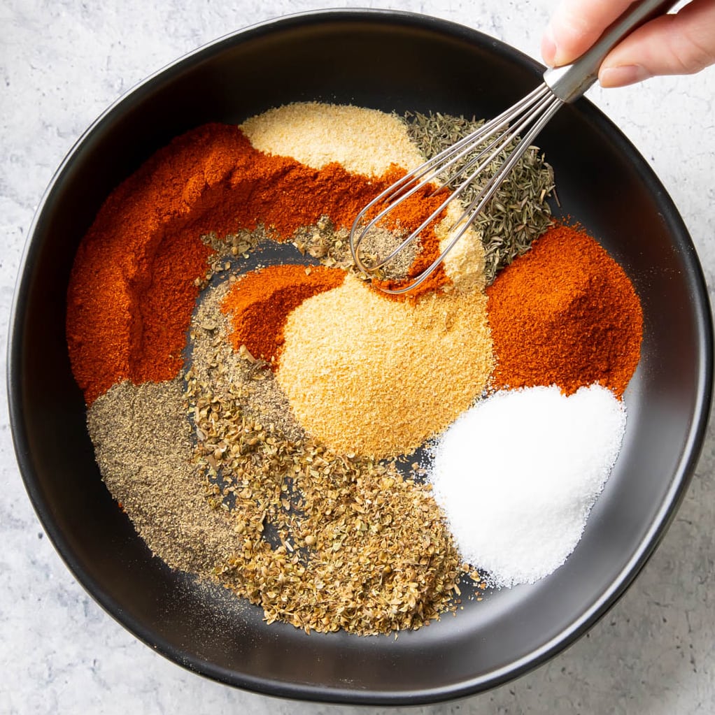 A hand stirring a small whisk into the dish of spices to combine them into Cajun Seasoning
