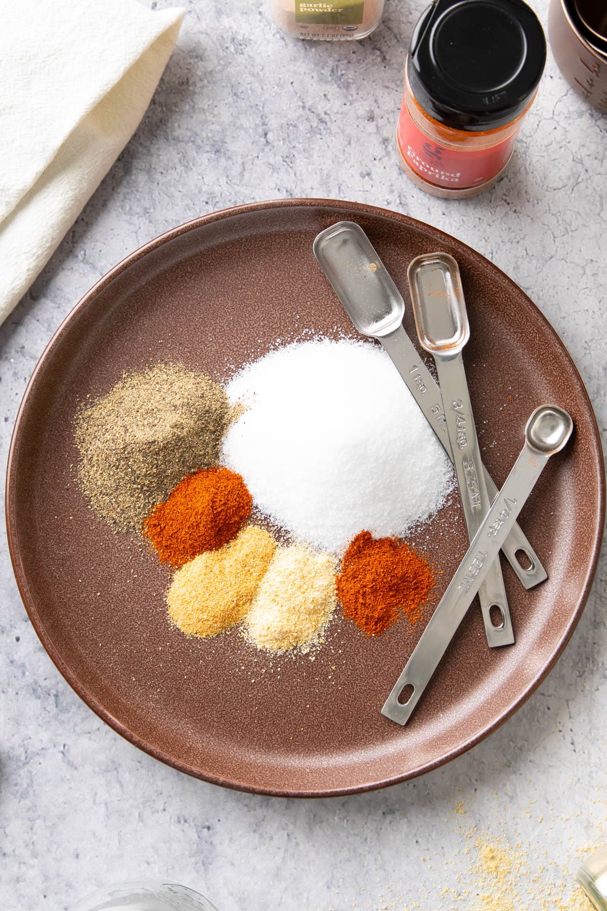 Seasoned salt spices and ingredients on a plate with measuring spoons, including salt, black pepper, paprika, cayenne, garlic, and onion powder.