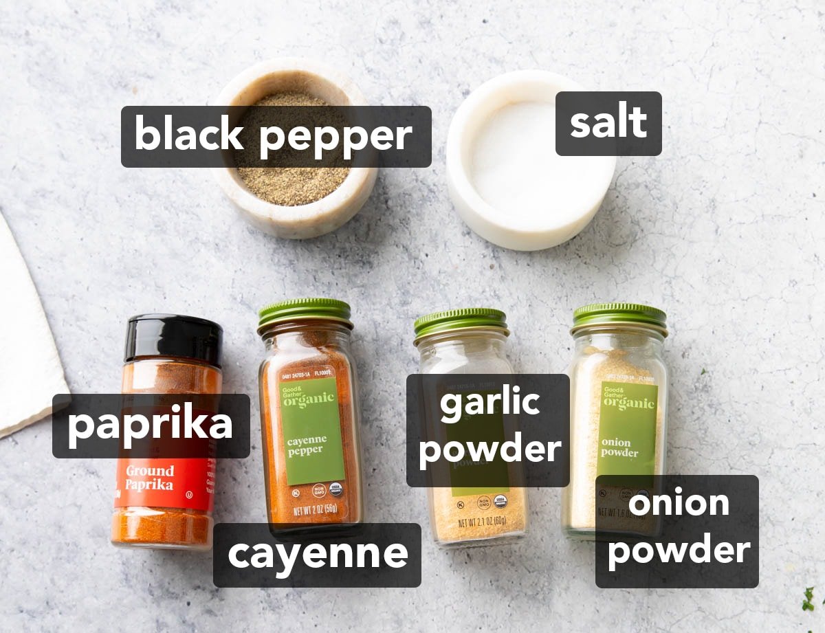 Seasoning salt ingredients on a table, including little bowls of ground black pepper and table salt, and labelled spice jars filled with ground paprika, cayenne pepper, garlic powder, and onion powder.