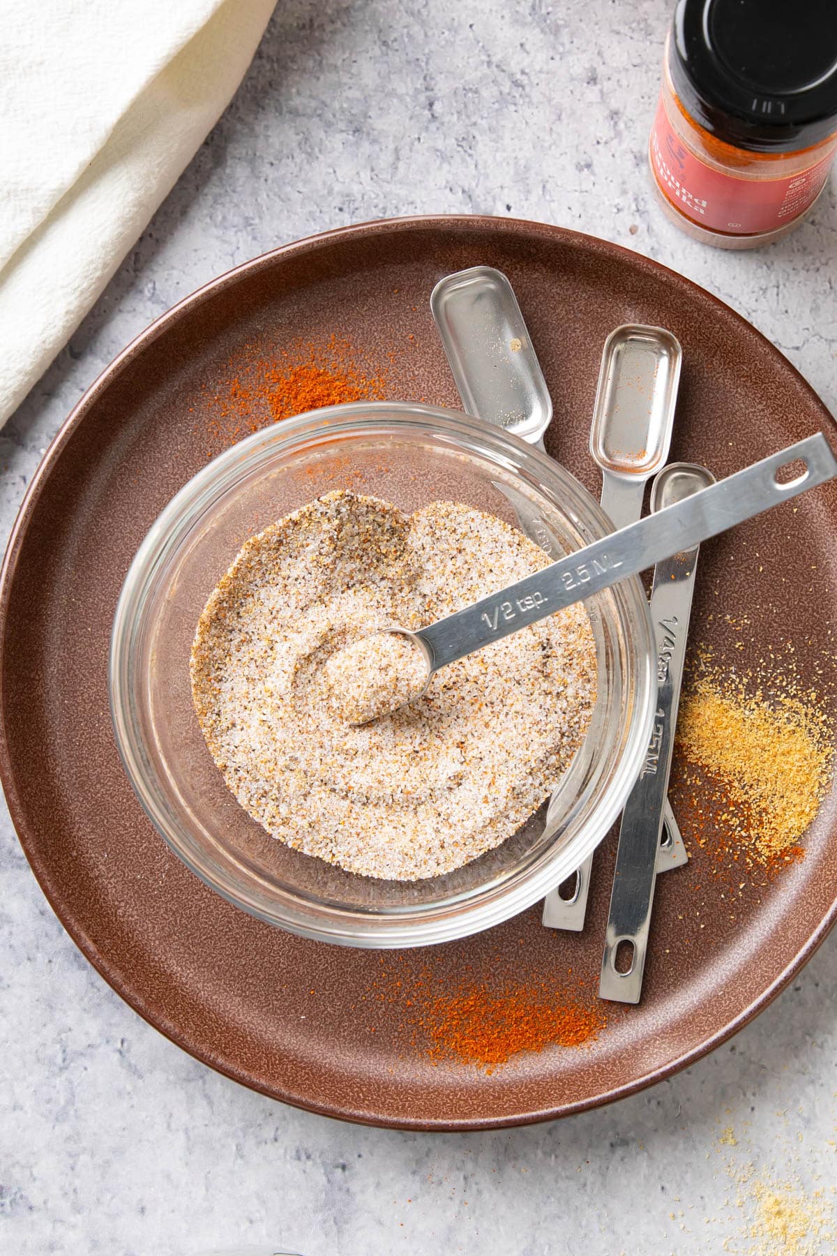 Measuring spoon sitting in a bowl of this homemade seasoning with bits of garlic powder, onion powder, cayenne, and more sprinkled around.