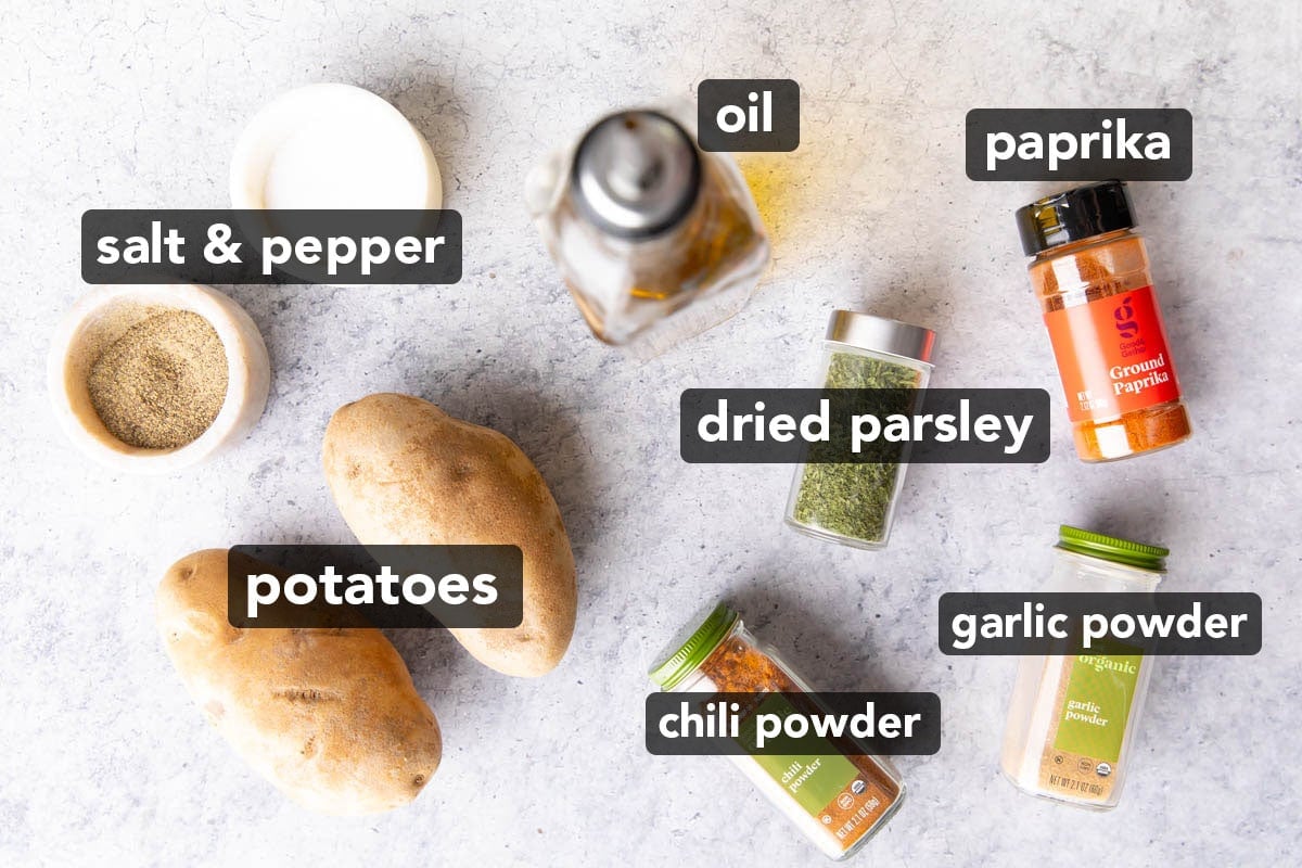 The ingredients for air fryer potato wedges laid out on a table including russet potatoes, oil, paprika, garlic powder, dried parsley flakes, chili powder, salt, and pepper.