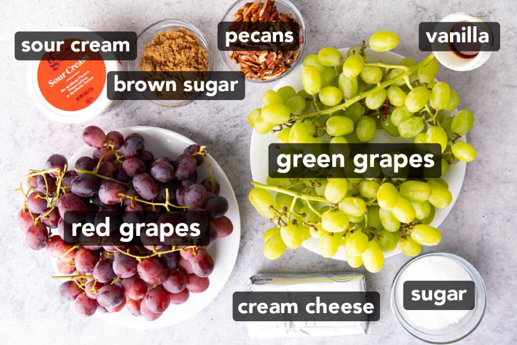 Grape Salad ingredients laid out on a table, including green grapes, red grapes, vanilla extract, sugar, cream cheese, pecans, brown sugar, and sour cream