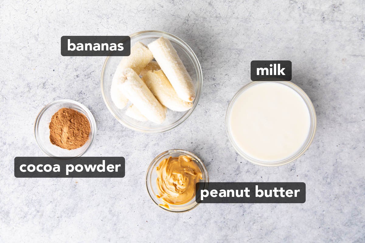 Ingredients for making this chocolate peanut butter banana smoothie in prep bowls, including cocoa powder, bananas, peanut butter, and milk