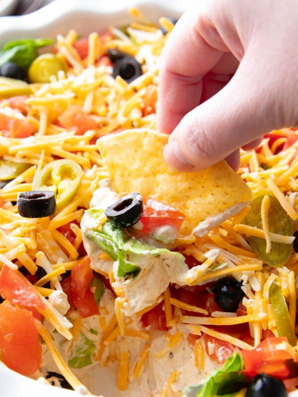 A hand dipping a tortilla chip into this layered taco dip and retrieving cream cheese seasoned dip with a black olive, lettuce, tomatoes, and cheese