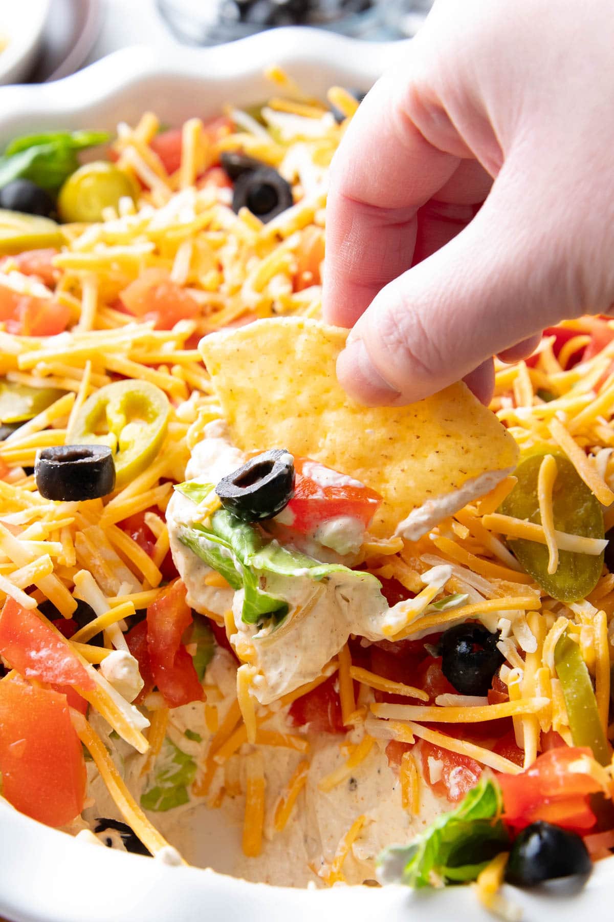 A hand dipping a tortilla chip into this layered taco dip and retrieving cream cheese seasoned dip with a black olive, lettuce, tomatoes, and cheese