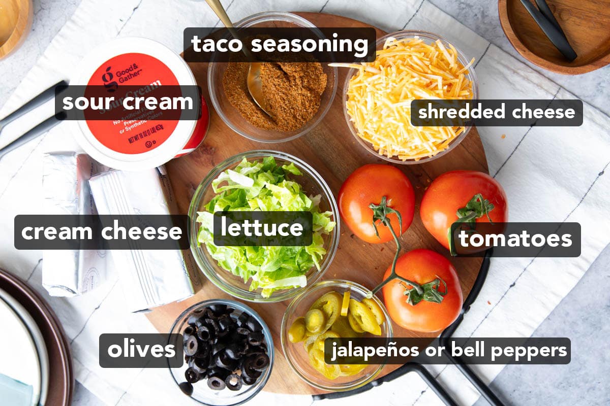 Easy Taco Dip ingredients laid out on a serving platter including homemade taco seasoning, sour cream, shredded cheese, tomatoes on the vine, sliced jalapeños, sliced olives, two blocks of cream cheese, and shredded lettuce