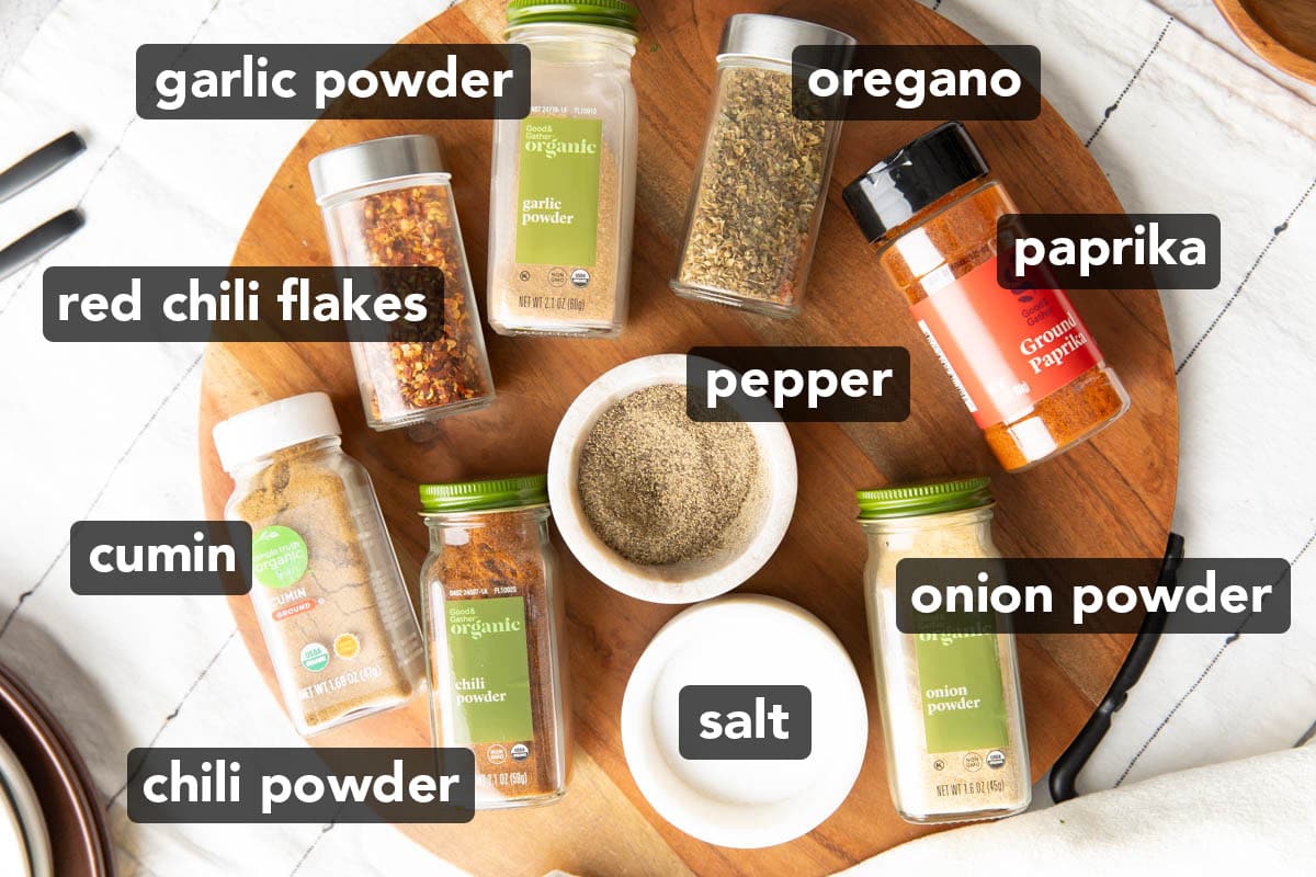 The ingredients for Taco Seasoning laid out on a table, including chili powder, ground cumin, garlic powder, paprika, salt, onion powder, dried oregano, black pepper, and red chili flakes.