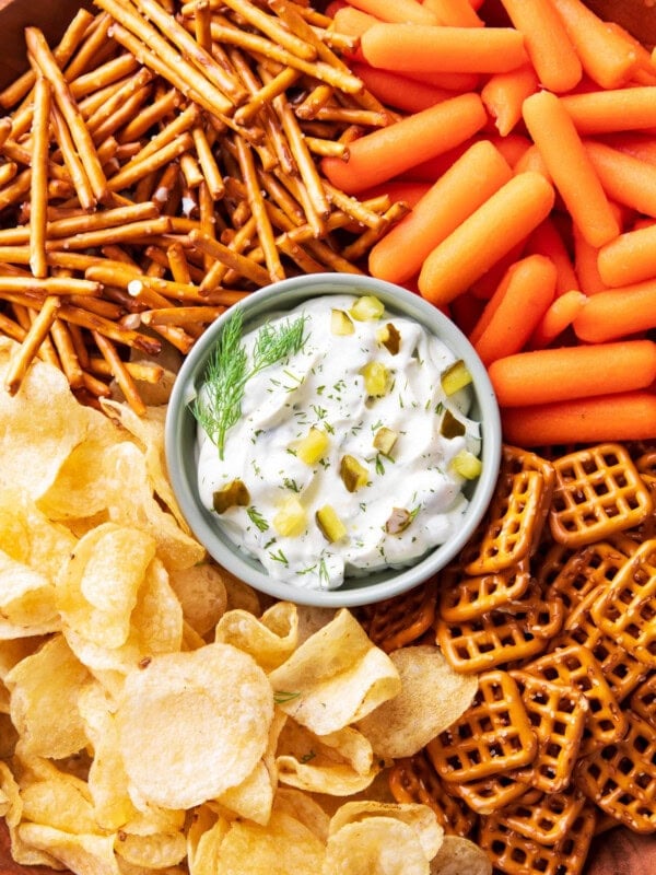 A serving platter with dill pickle dip in the center served with carrots, pretzels, and potato chips for dipping.