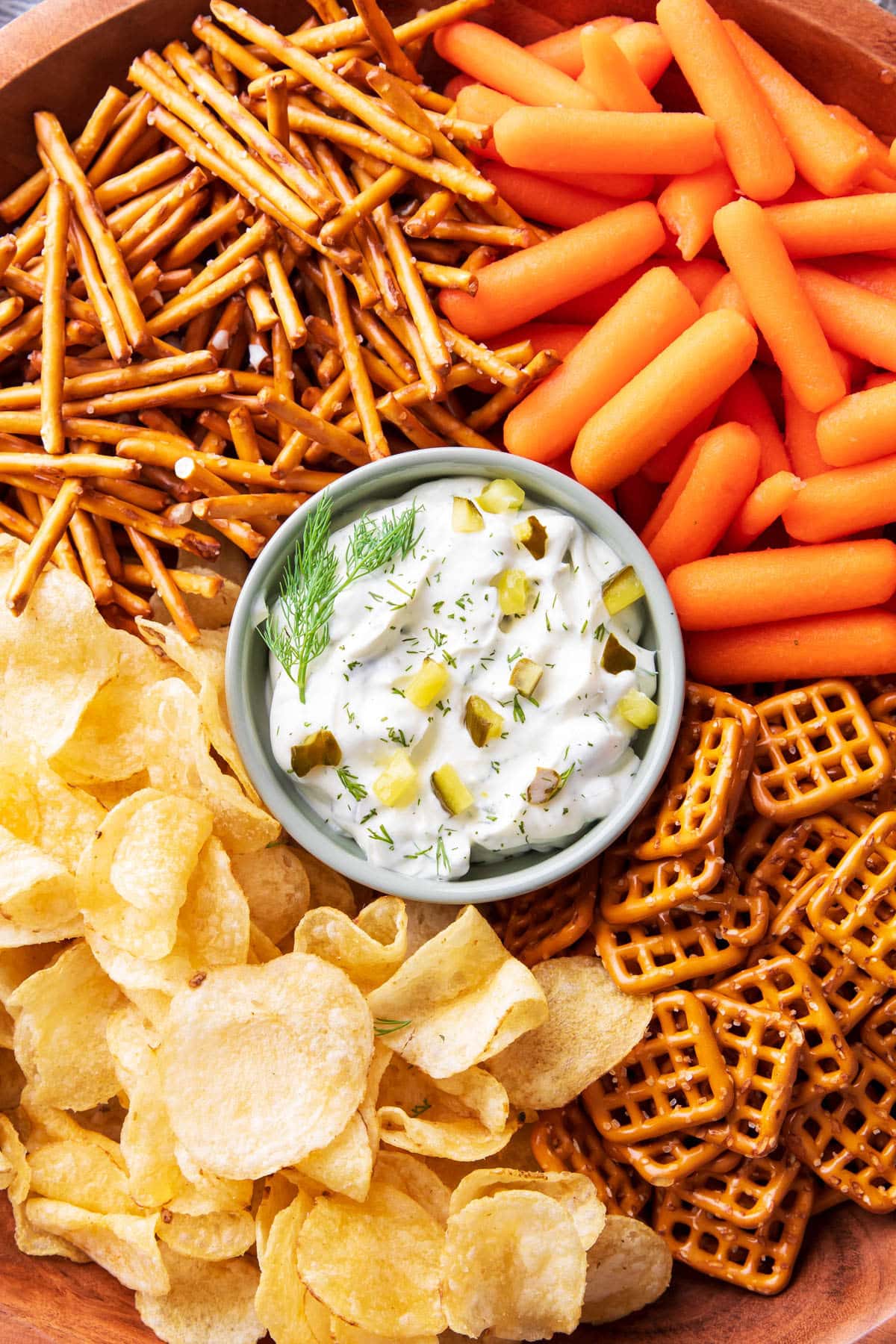 A serving platter with dill pickle dip in the center served with carrots, pretzels, and potato chips for dipping