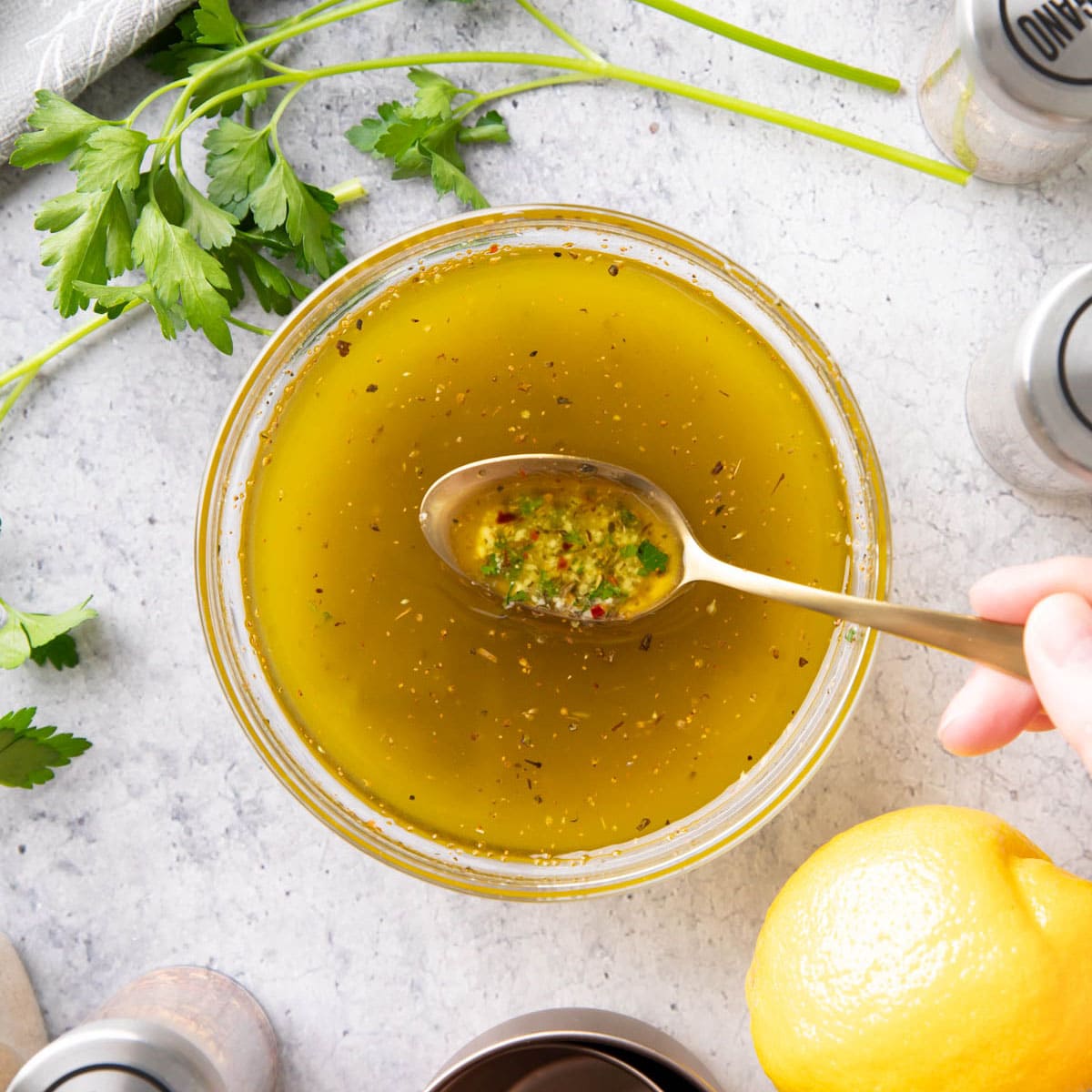 Spooning this Italian Dressing Recipe from a mixing bowl to showcase the ingredients, including herbs, olive oil, lemon juice, and more.