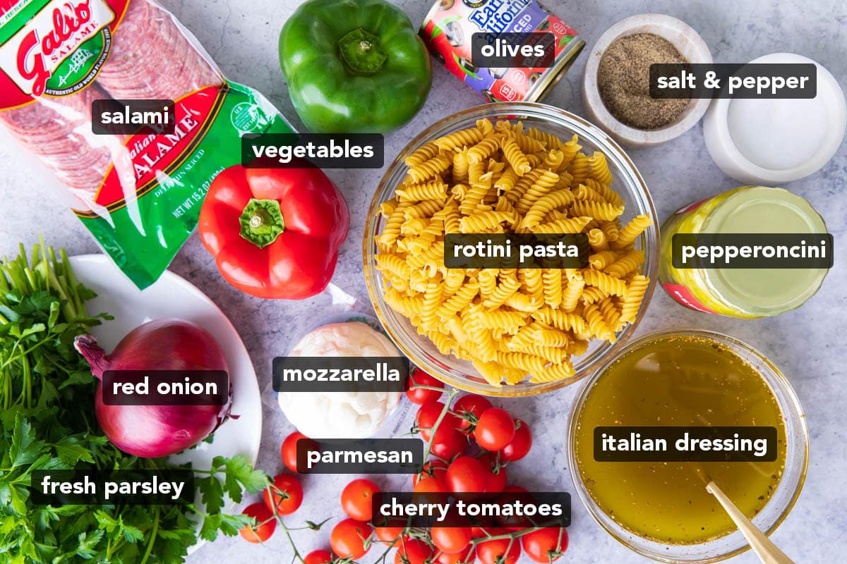 Ingredients for this Italian pasta salad recipe including pasta, salami, red onion, bell peppers, olives, tomatoes, mozzarella, pepperoncini, parsley and Italian dressing.