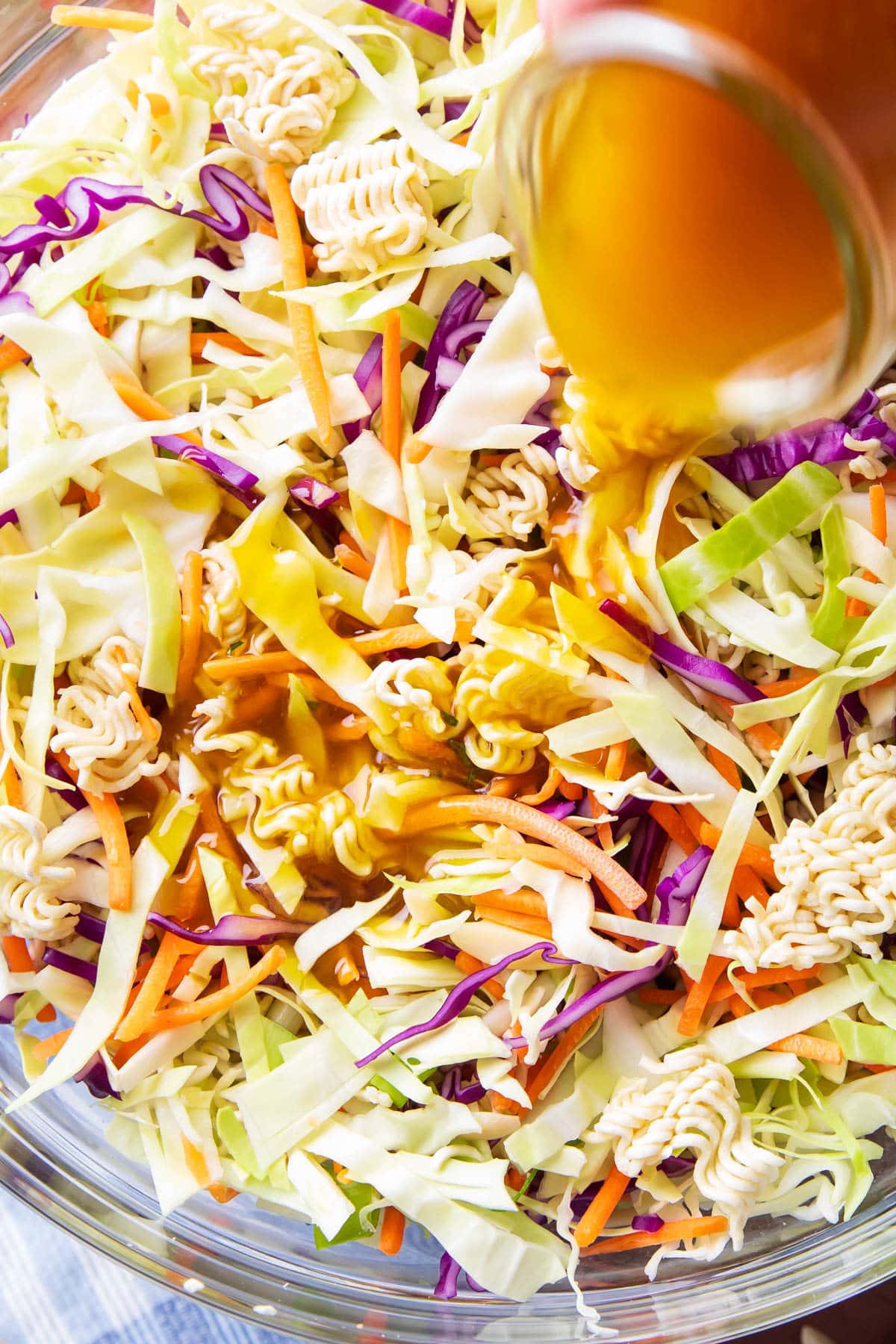 Pouring a jar of homemade dressing over ramen noodle salad packed with cabbage, green onions, almonds, and carrots.