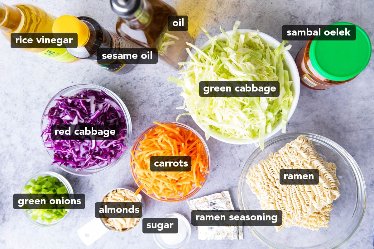 Ingredients for making ramen noodle salad recipe laid out on a table including sesame oil, ramen seasoning, uncooked ramen, red cabbage, carrots, and more.