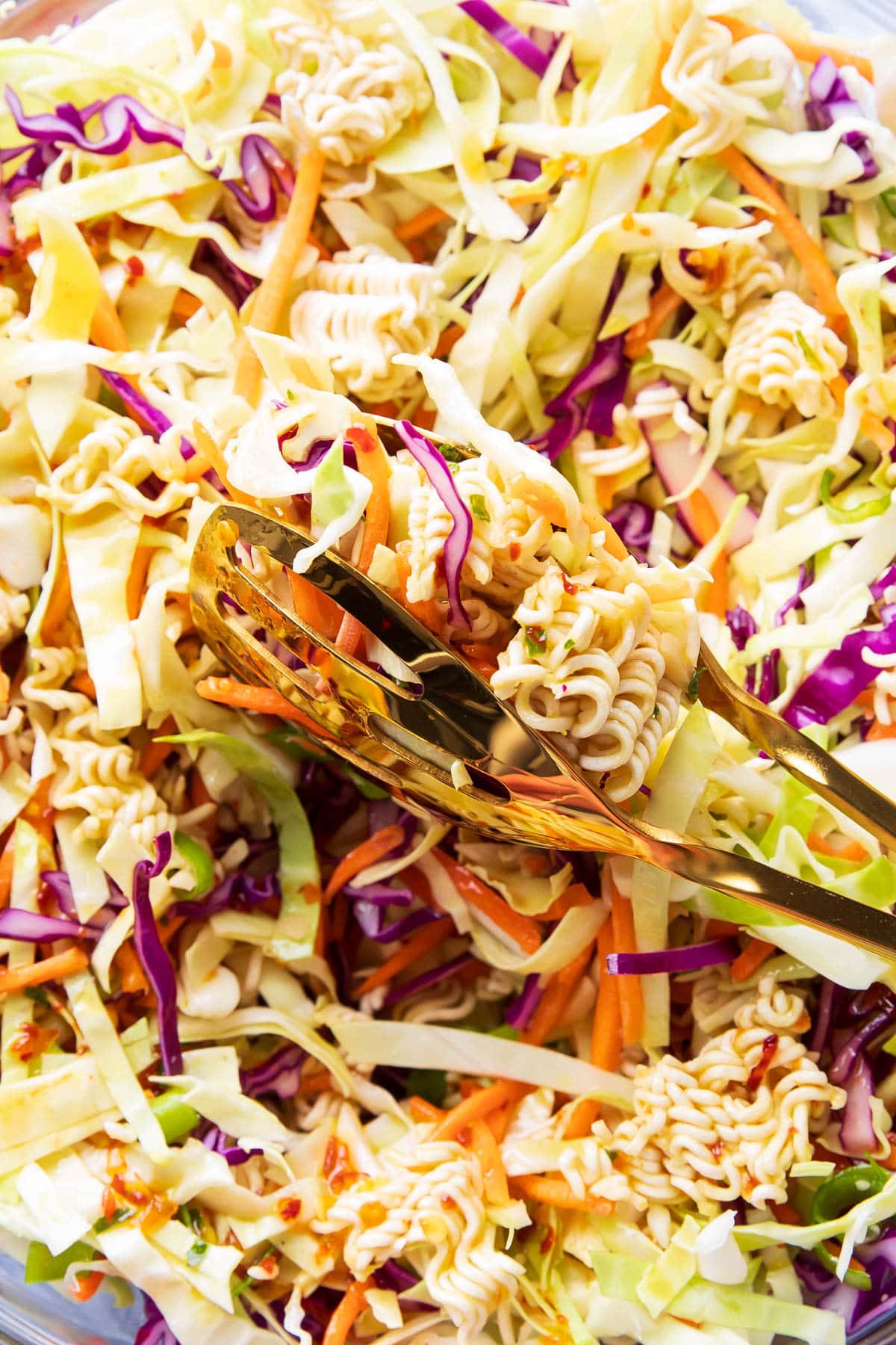 A pair of gold tongs holding up crunchy toppings and heaps of saucy asian vegetables