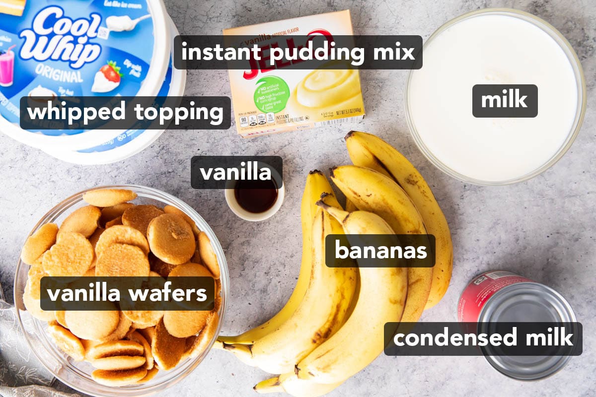 Southern banana pudding recipe ingredients laid out on a table including bananas, condensed milk, instant pudding mix, vanilla wafers, Cool Whip frozen topping, and vanilla extract.