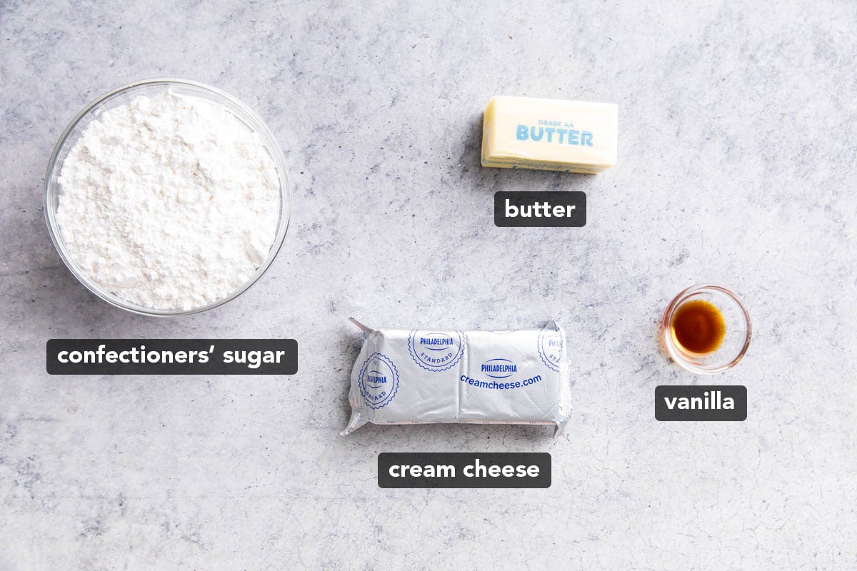 Cream cheese frosting ingredients laid out on a table including brick of cream cheese, confectioners’ sugar, butter, and vanilla extract