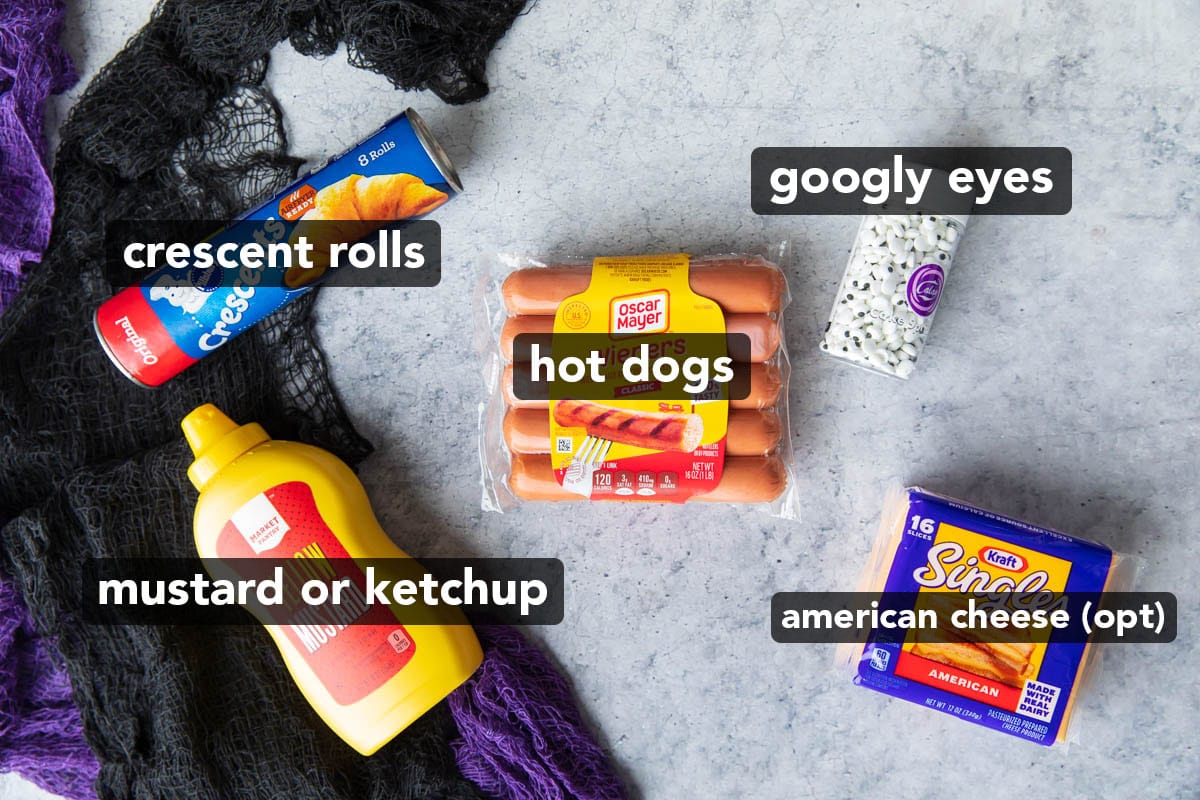 mummy hot dog recipe ingredients laid out on a table featuring hot dogs, mustard, crescent roll dough, googly eye sprinkles, and American cheese