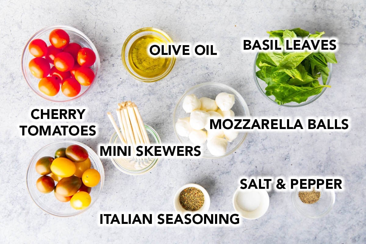 The ingredients for the best fancy caprese skewers recipe in prep bowls including tomatoes, olive oil, mozzarella balls, seasoning, mini skewers, fresh basil leaves, salt and pepper.