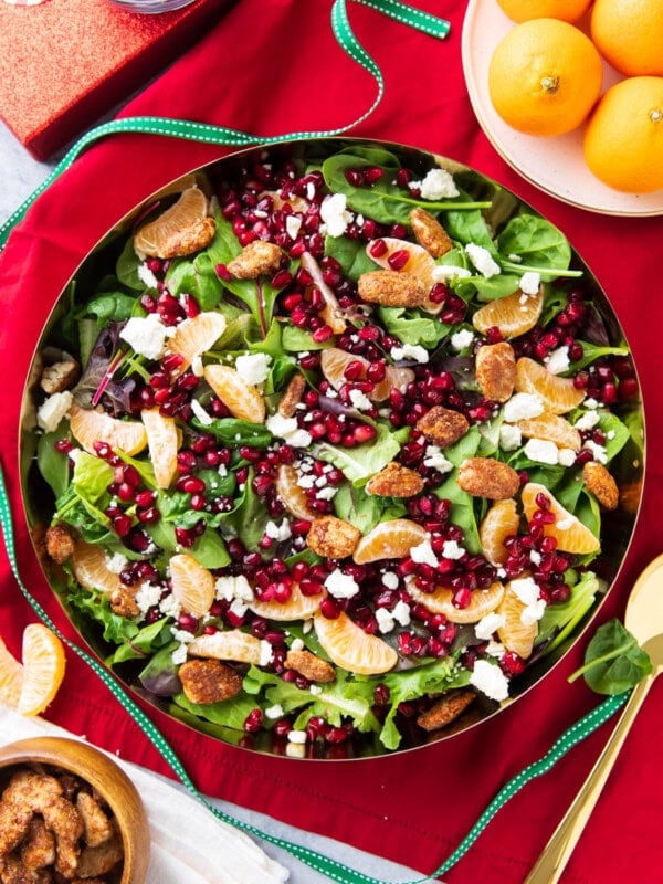 Christmas Salad packed full of baby lettuce, arugula, spinach, oranges, nuts, and cheese served in a large salad bowl with festive red table cloth.