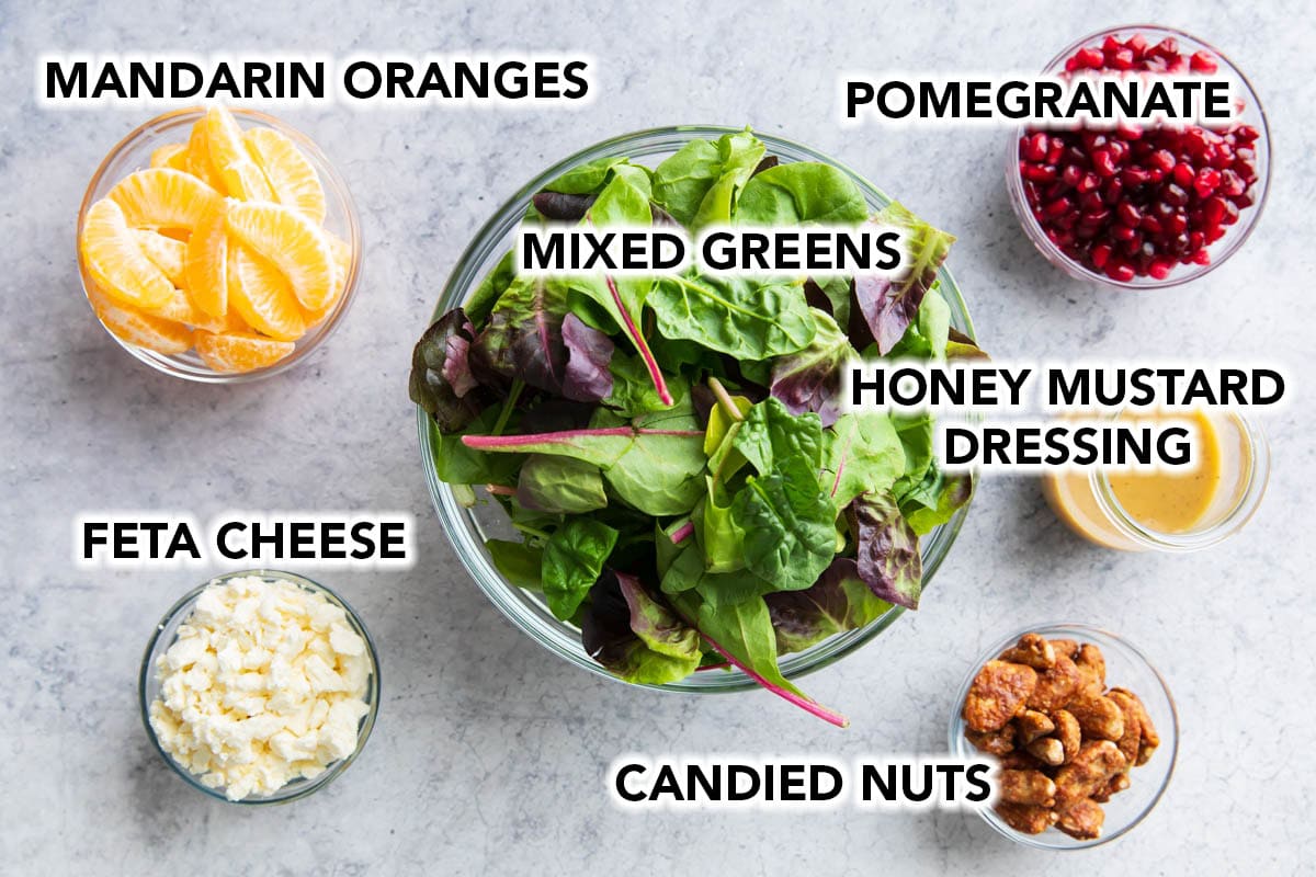 The ingredients for this Christmas Salad recipe in prep bowls, including candied nuts, pomegranates, spring mix green, honey mustard dressing, mandarin oranges, and feta cheese.