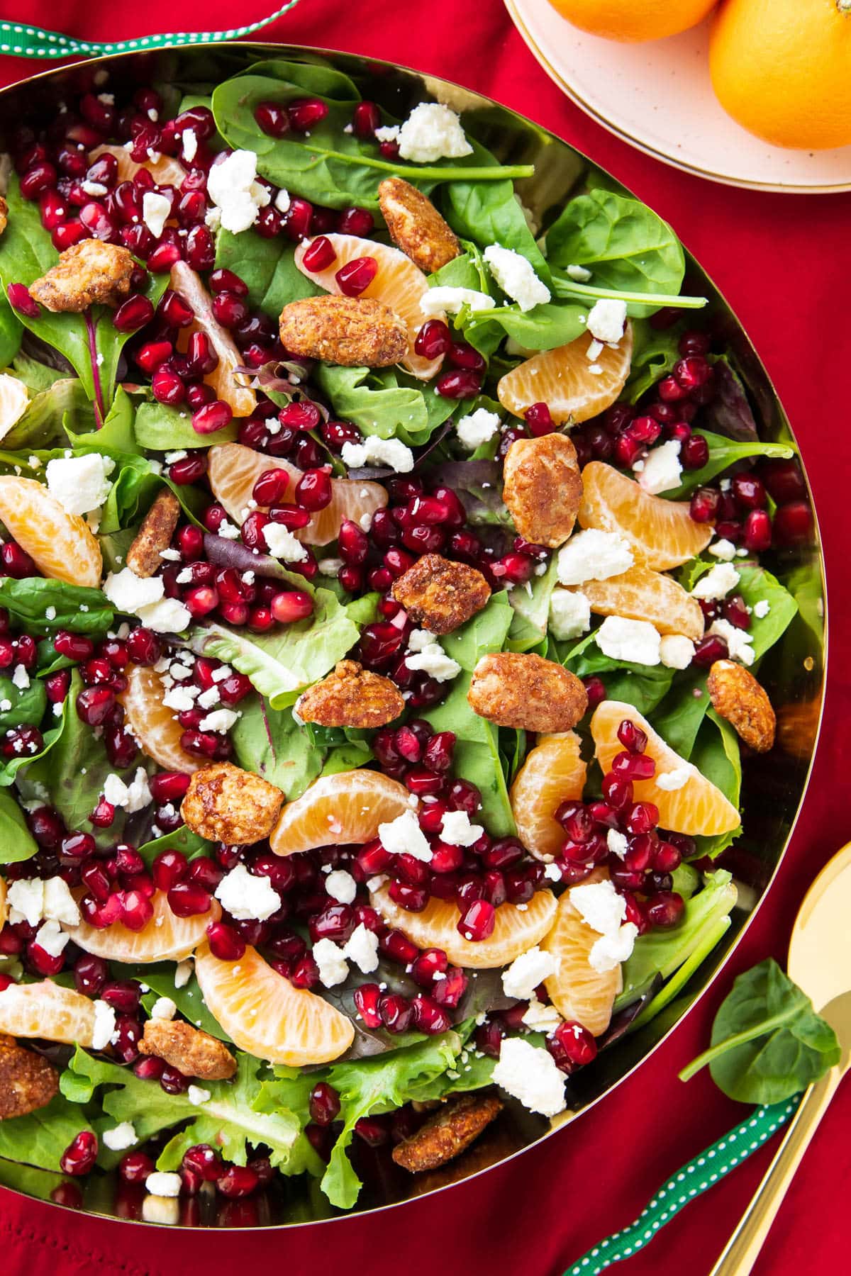 Christmas Salad packed full of baby lettuce, arugula, spinach, oranges, nuts, and cheese served in a large salad bowl with festive red table cloth.