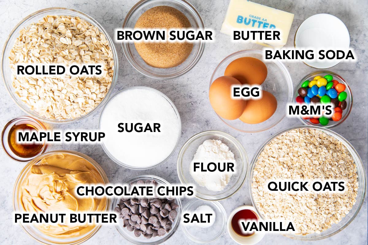 The ultimate monster cookie recipe ingredients prepped in bowls including rolled oats, quick oats, flour, brown sugar, vanilla extract, peanut butter, eggs, butter, chocolate chips, and candies.