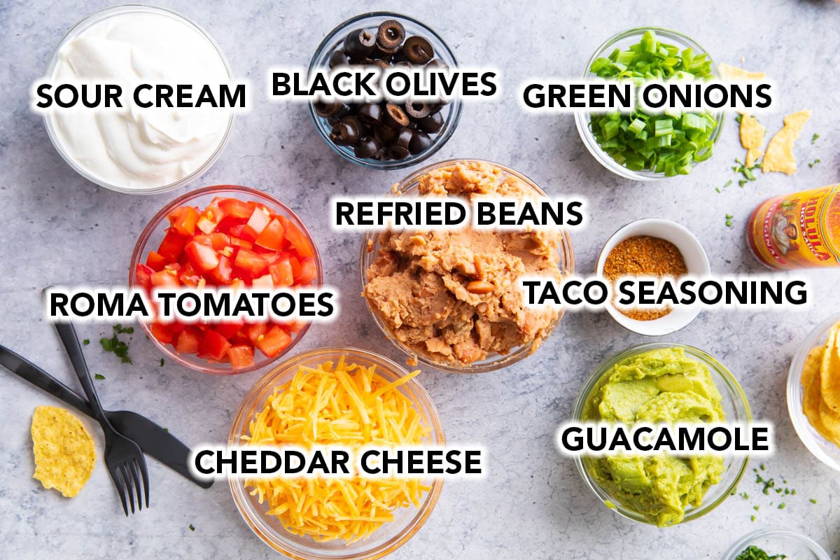 the ingredients for this old fashioned 7 layer dip including refried beans, guacamole, black olives, tomatoes, cheese, green onions, taco seasoning, and sour cream.