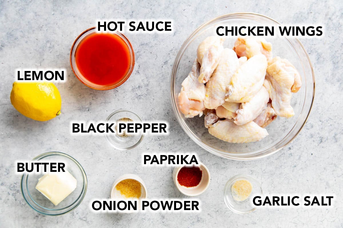 Ingredients for making buffalo wings in air fryer including raw chicken wings, hot sauce, butter, lemon, and spices.