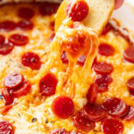 Someone dipping a bagel chip into pizza dip, stretching pepperonis and cheese from the dish.