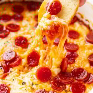 Someone dipping a bagel chip into pizza dip, stretching pepperonis and cheese from the dish.