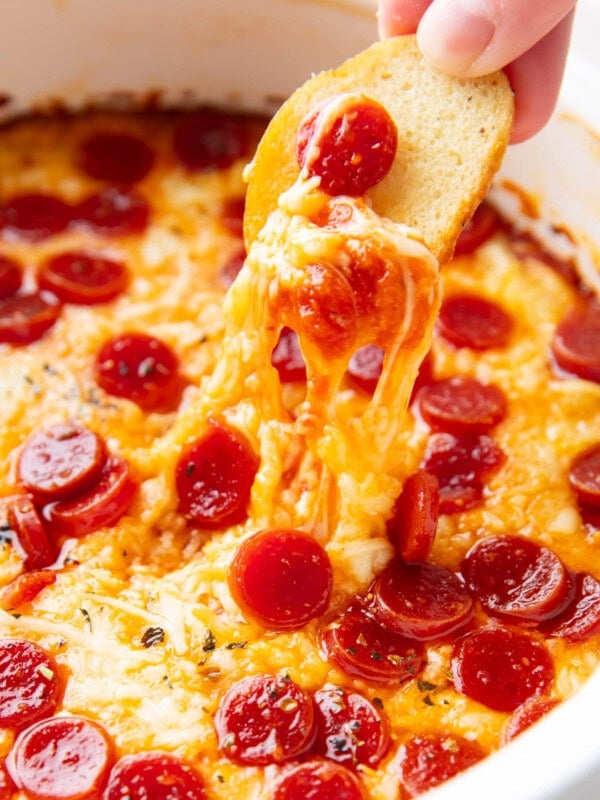 Someone dipping a bagel chip into pizza dip, stretching pepperonis and cheese from the dish.