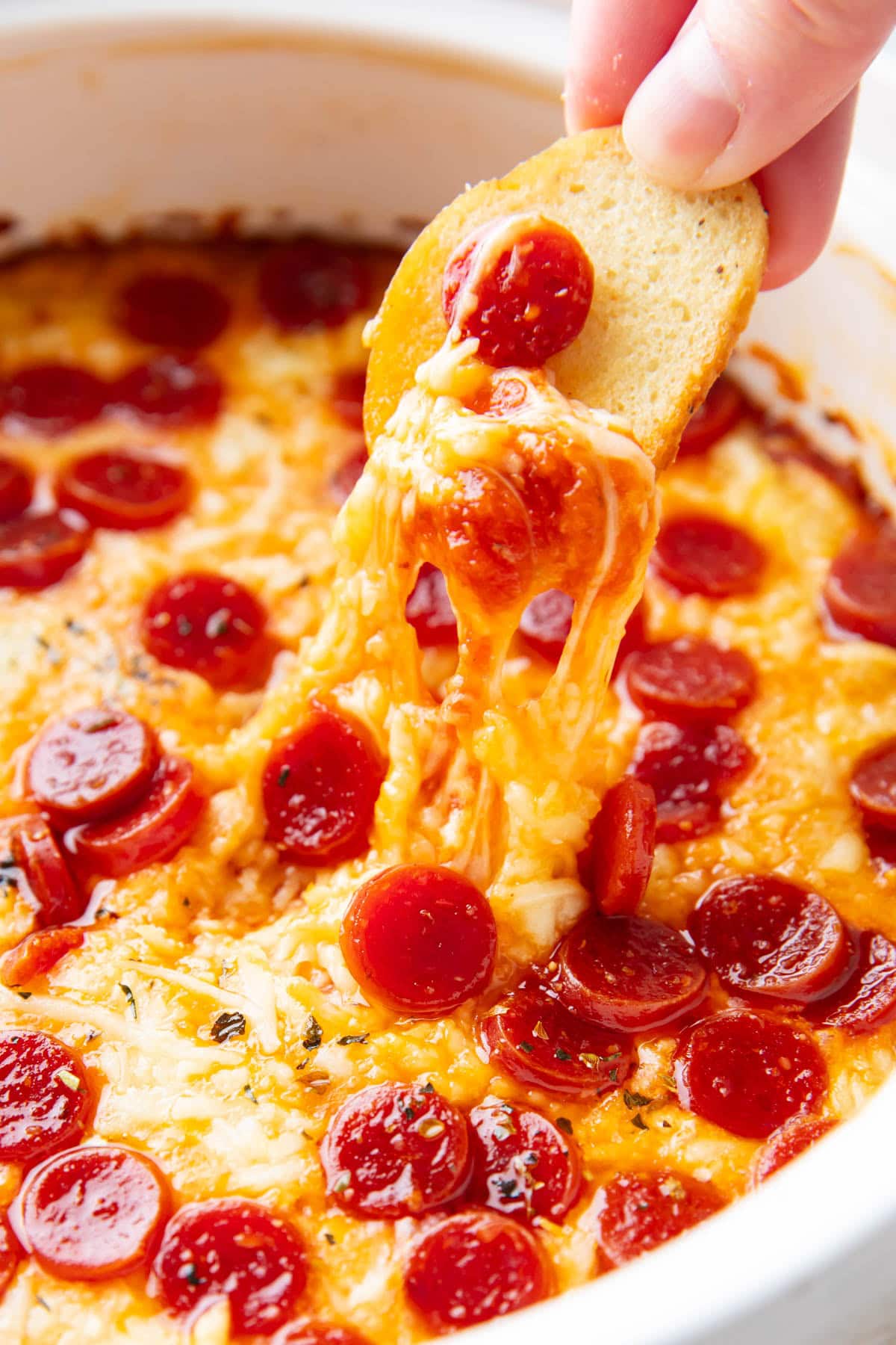 Someone dipping a bagel chip into pizza dip, stretching pepperonis and cheese from the dish.