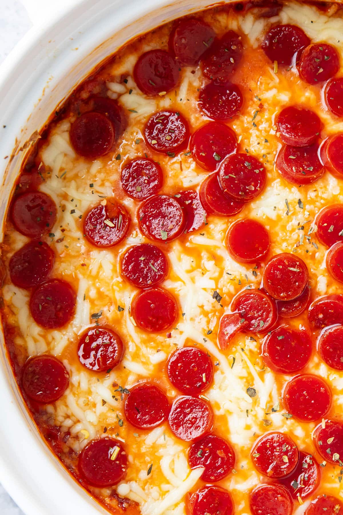 Overhead photo of hot pizza dip with greasy pepperoni, melted cheese, and seasonings in a casserole dish.