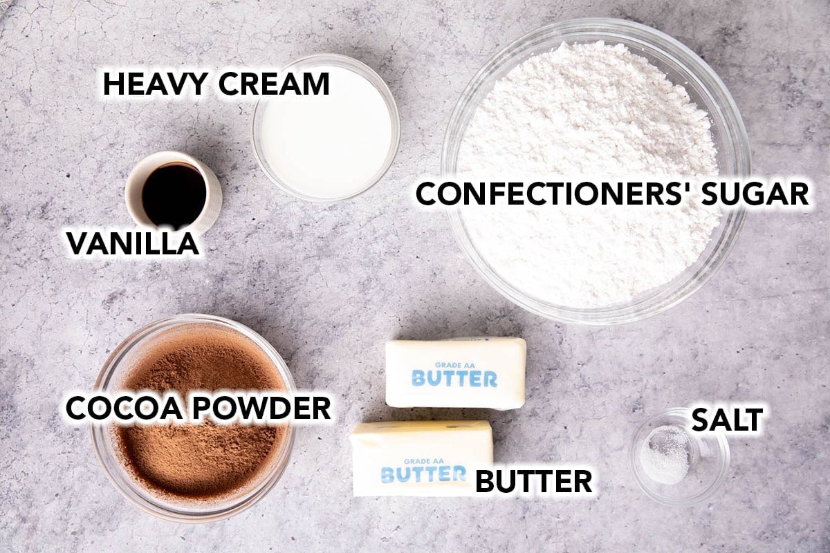 chocolate frosting recipe ingredients on a table including confectioners sugar, butter, cocoa powder, vanilla extract, and heavy cream