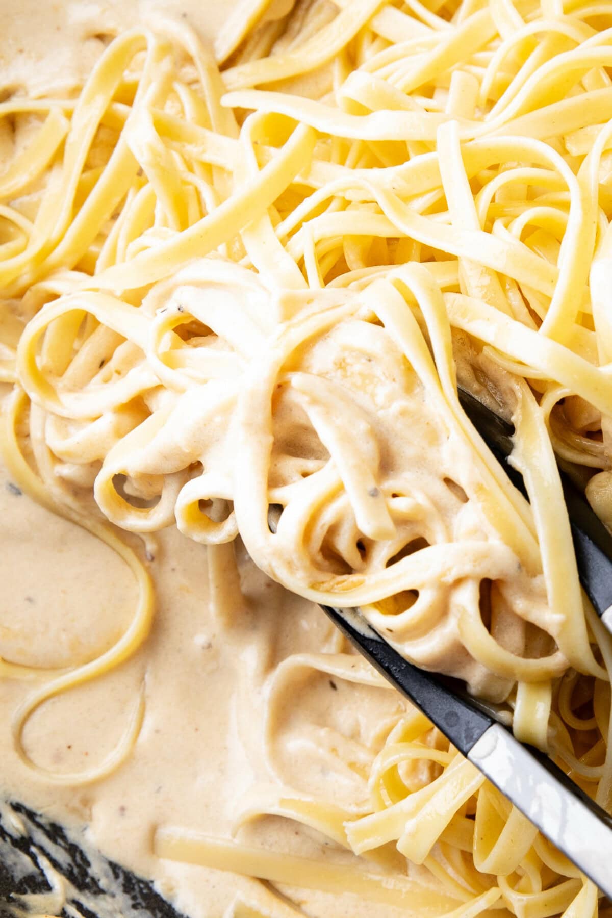 close up of this Fettuccine Alfredo dish - tongs hold freshly cooked fettuccine pasta to fold in creamy homemade Alfredo sauce