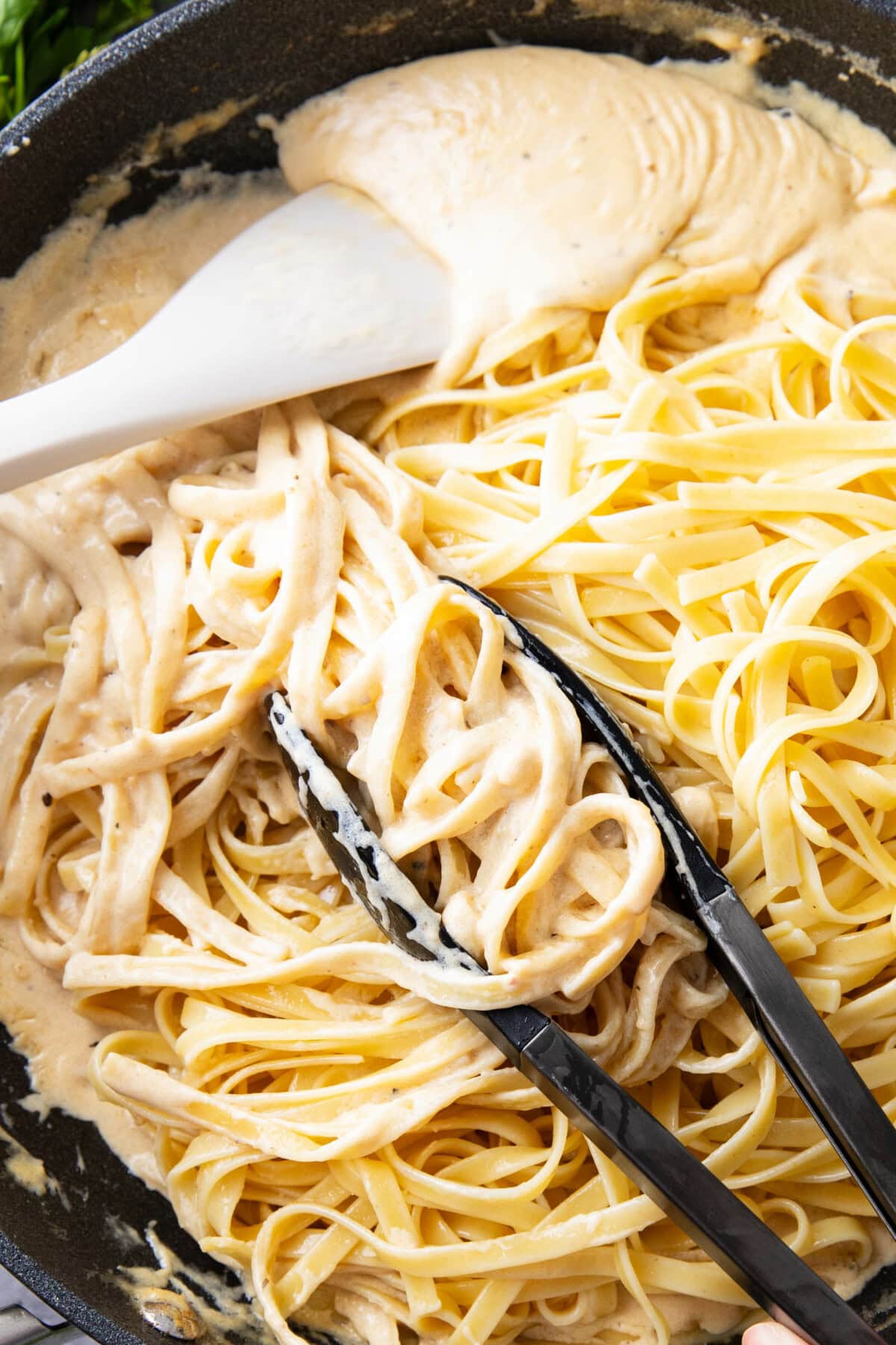 tongs grabbing cooked fettuccine and tossing in Alfredo sauce with spatula in homemade Alfredo sauce