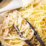 tongs grabbing cooked fettuccine and tossing in Alfredo sauce with spatula in homemade Alfredo sauce